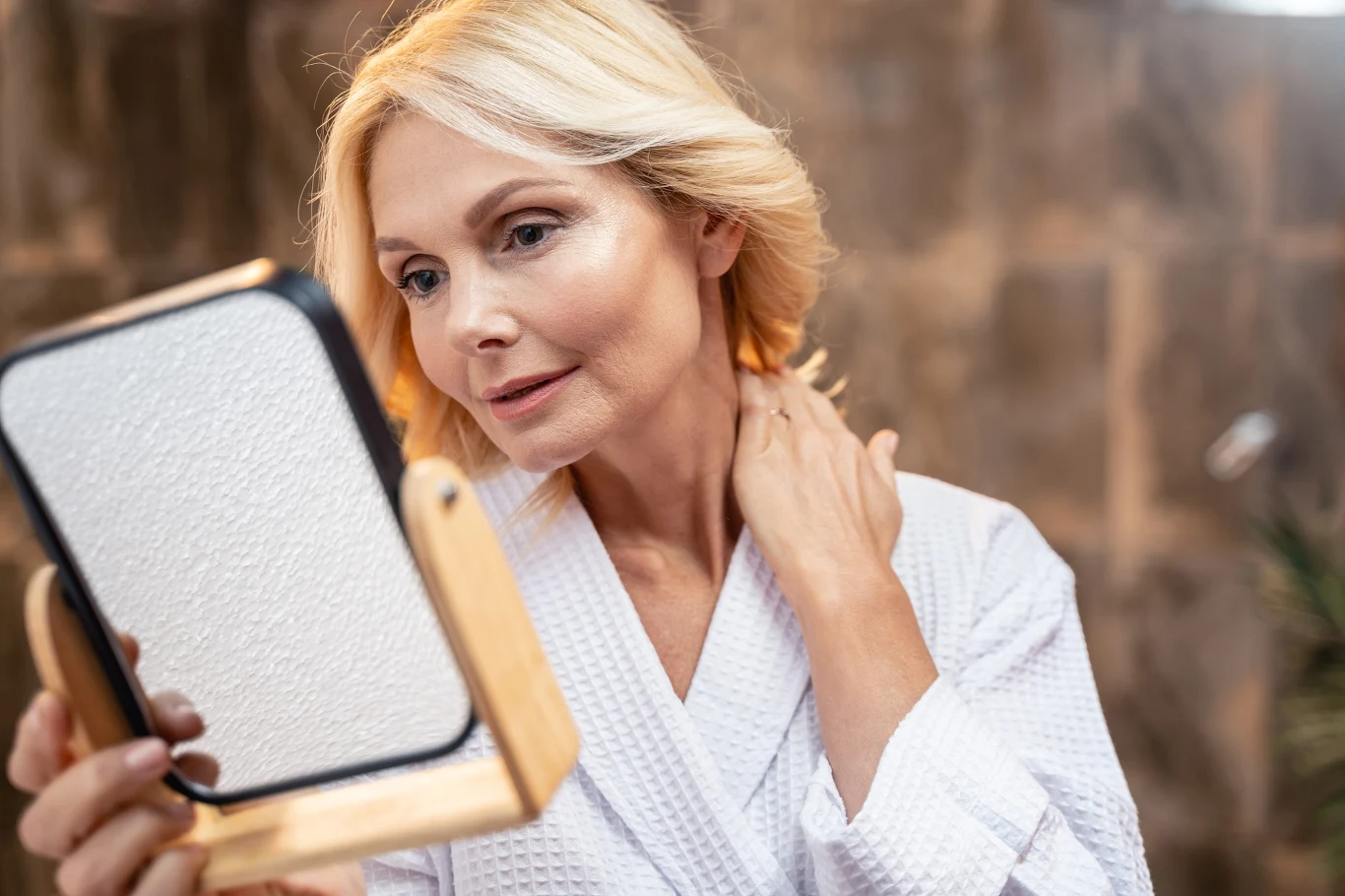 Po 40. wcale nie trzeba wprowadzać agresywnej pielęgnacji "anti aging" Kobieta w jasnym szlafroku trzyma lusterko i dotyka szyi, przyglądając się swojej twarzy.