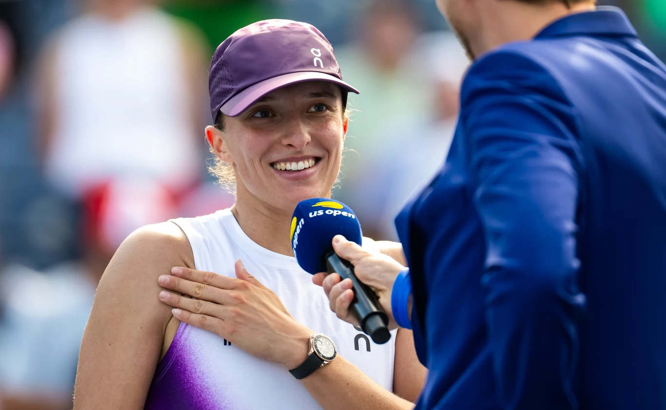 Uśmiechnięta tenisistka w czapce i sportowej koszulce podczas wywiadu na korcie, trzymająca rękę na ramieniu, obok osoba z mikrofonem US Open.
