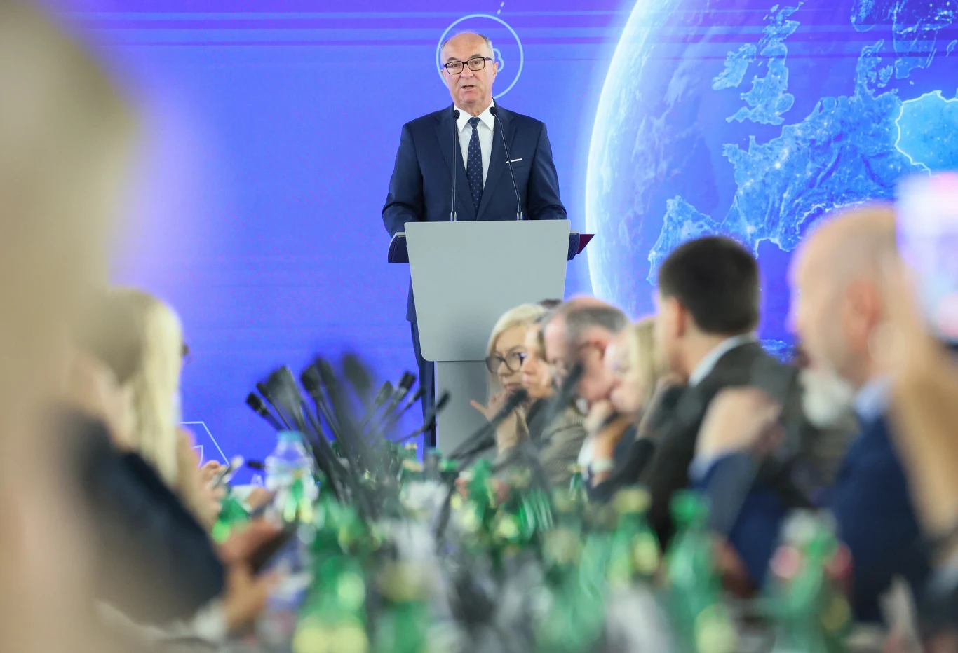 Mężczyzna w garniturze przemawia przy mównicy na tle mapy Europy, a przed nim zasiadają uczestnicy spotkania przy stole z butelkami wody i mikrofonami.