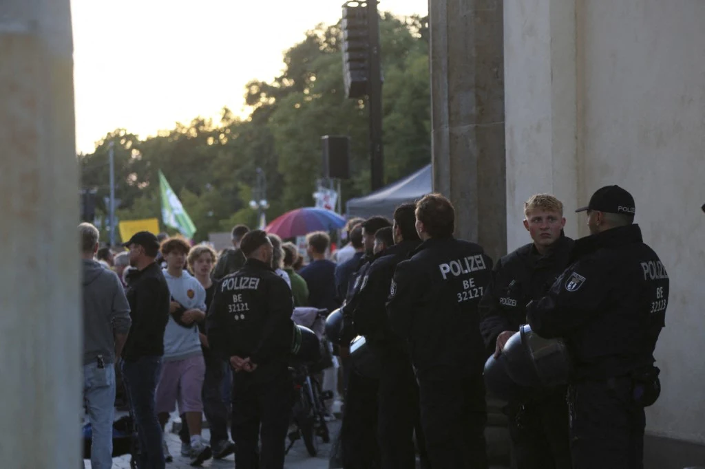 Grupa policjantów w ciemnych mundurach obserwujących tłum ludzi zgromadzonych na ulicy, w tle widoczne flagi i namioty, atmosfera sugerująca wydarzenie publiczne lub demonstrację.