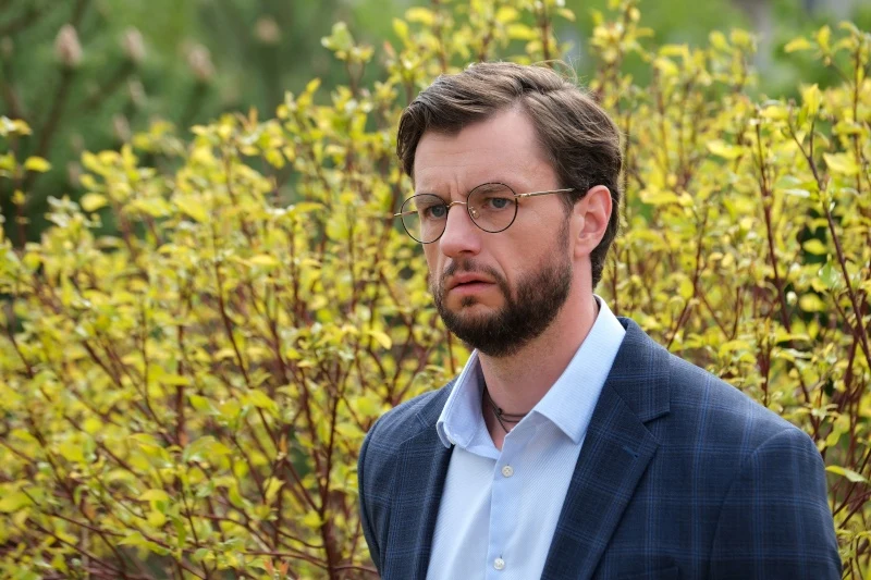 Mariusz zobaczy Kasię z Sowińskim Mężczyzna z brodą i okularami, ubrany w niebieską marynarkę i jasną koszulę, stoi na tle krzewów z żółtymi liśćmi.