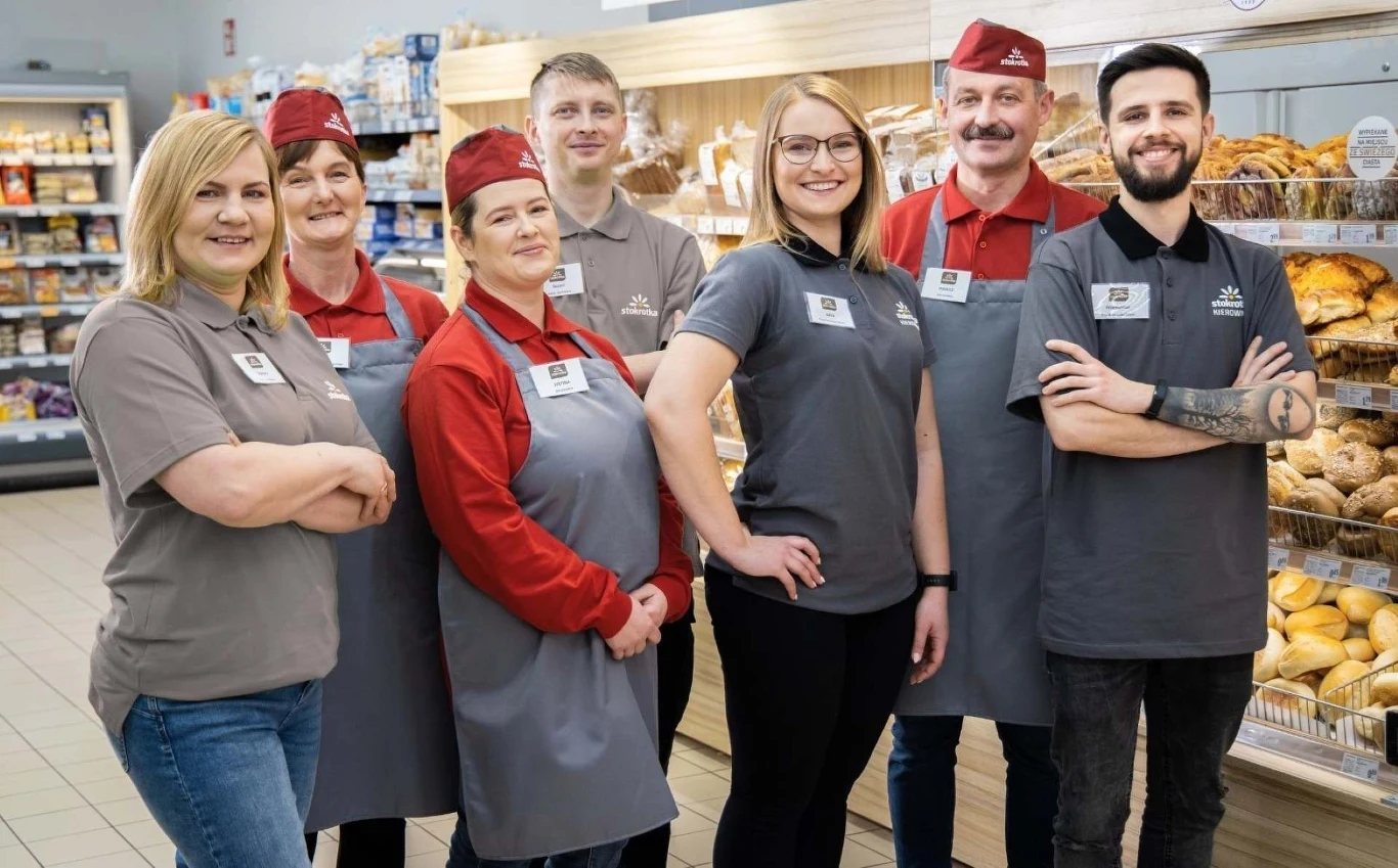 Stokrotka Grupa pracowników sklepu spożywczego w uniformach stoi uśmiechnięta przed półką z pieczywem.