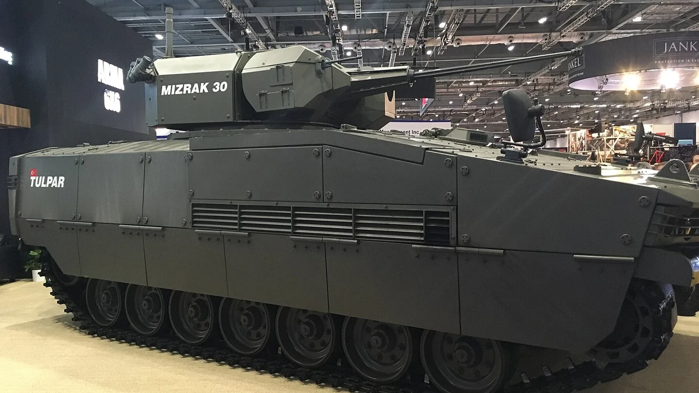 Opancerzony pojazd gąsienicowy typu Tulpar uzbrojony w wieżę MIZRAK 30, zaprezentowany w hali wystawowej pod sztucznym oświetleniem.