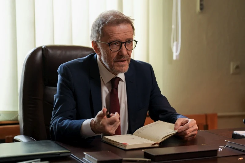 Prokurator zapyta Marię, dlaczego tak długo milczała Starszy mężczyzna w okularach i garniturze siedzi przy biurku z otwartym notesem w dłoni, wykonując gest dłonią podczas rozmowy.