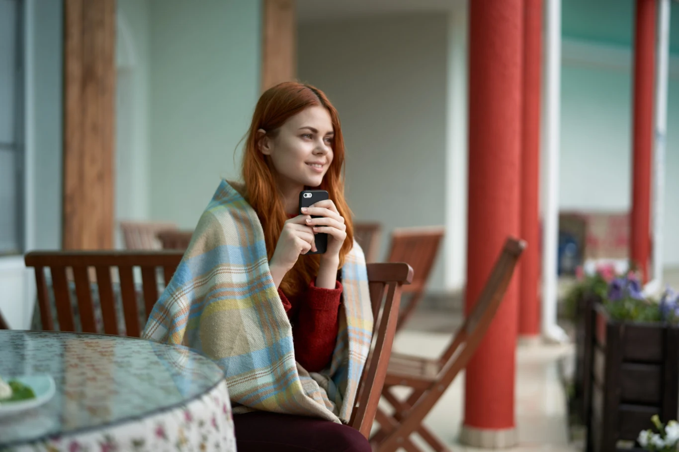 Młoda kobieta owinięta kocem siedzi przy stole na tarasie, trzymając w dłoniach kubek i telefon, z delikatnym uśmiechem spogląda w dal.