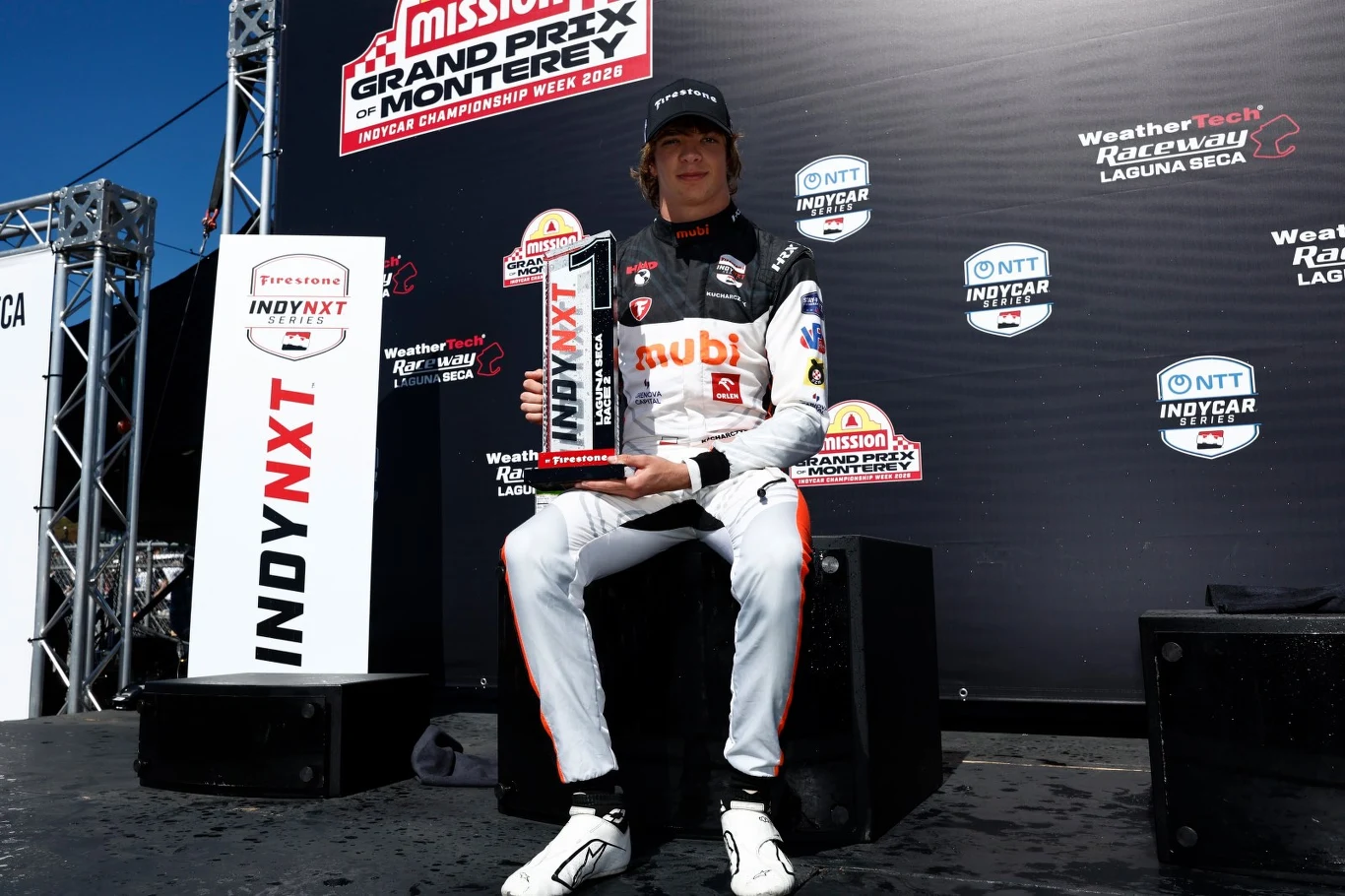 Kierowca wyścigowy w białym kombinezonie z czarnymi i czerwonymi detalami siedzi na podium, trzymając puchar za zwycięstwo podczas wyścigu Grand Prix Monterey, za nim widoczne są banery sponsorów i logo toru wyścigowego.