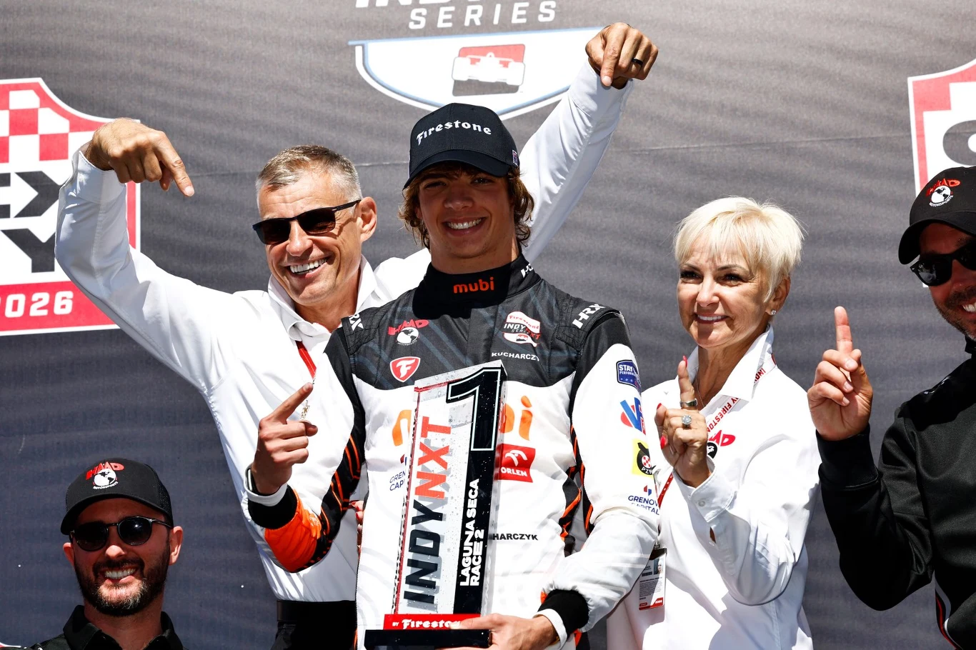 Zwycięzca wyścigu Indy NXT trzyma duże trofeum z numerem 1, uśmiecha się i otoczony jest przez zespół, który wskazuje na niego, świętując wygraną.