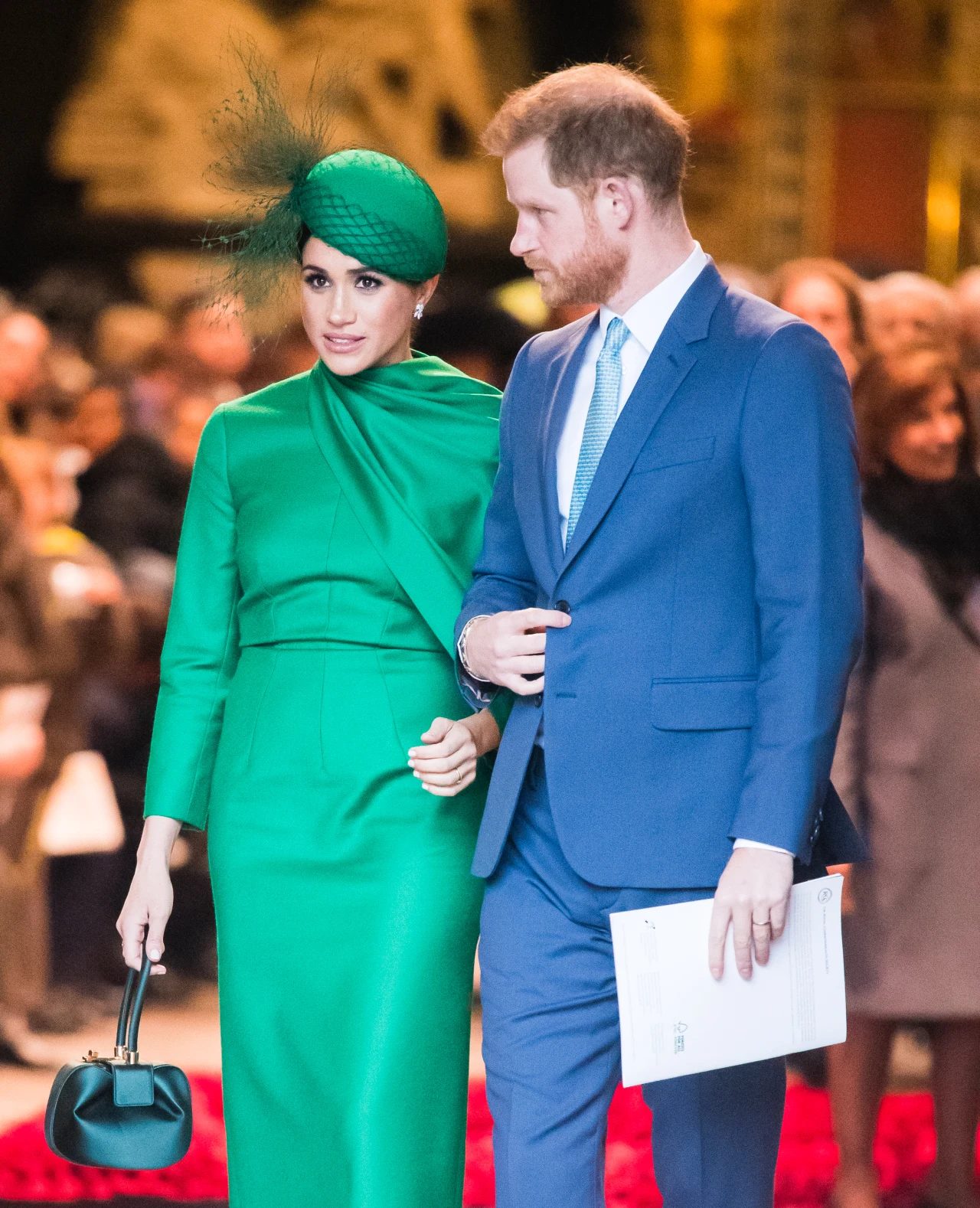 Elegancka para w centrum uwagi — kobieta w intensywnie zielonej sukni z dopasowanym fascynatorem i torebką, mężczyzna ubrany w niebieski garnitur z białą koszulą i jasnoniebieskim krawatem, idą razem w uroczystym otoczeniu.