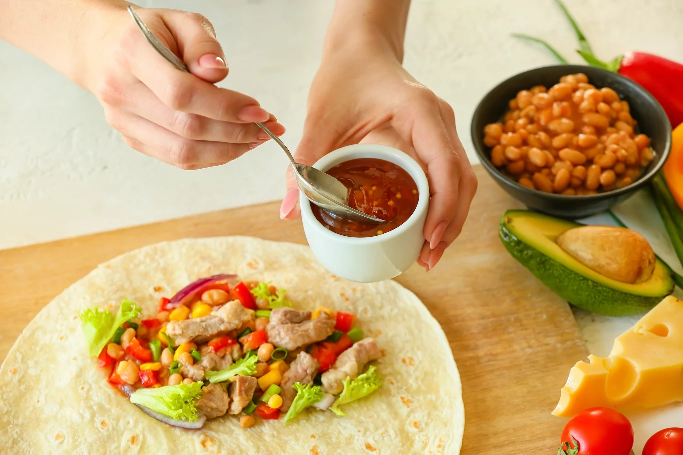 Do tortilli możesz włożyć ulubione dodatki. Jeśli nie chcesz, aby była kaloryczna, zrezygnuj z majonezu i tłustych sosów Tortilla z kawałkami mięsa, sałatą, kukurydzą, papryką i cebulą, do której dodawany jest sos pomidorowy; obok widoczne awokado, starty ser, fasola i warzywa.