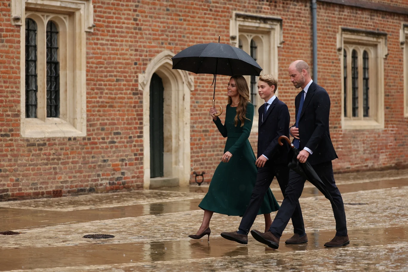 Trzy osoby elegancko ubrane przechodzą brukowaną ulicą w deszczowy dzień, jedna z nich trzyma czarny parasol nad grupą, w tle ceglany budynek z dużymi oknami.