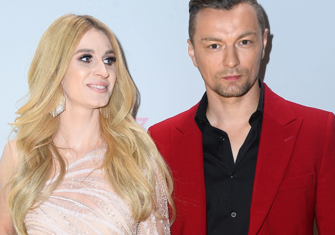 Kobieta z długimi blond włosami w eleganckiej, jasnej sukni i mężczyzna w czerwonej marynarce oraz czarnej koszuli stoją obok siebie, patrząc w różne strony.