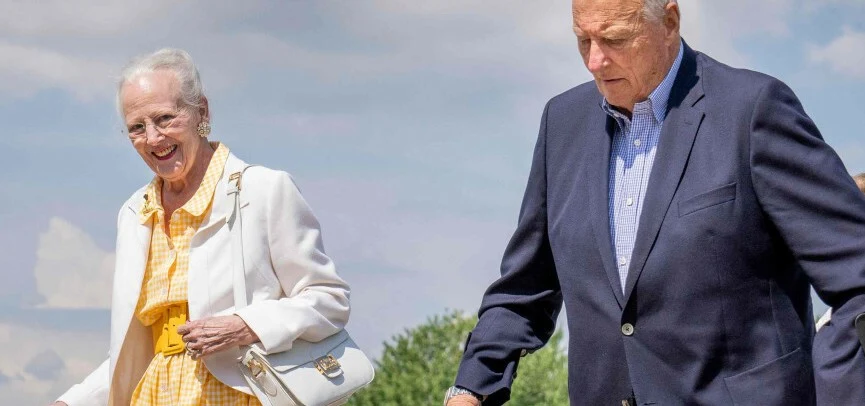 Starsza kobieta w okularach ubrana w żółtą sukienkę i białą marynarkę idzie obok starszego mężczyzny w granatowej marynarce i koszuli, oboje na tle jasnego, słonecznego nieba.