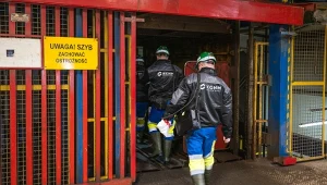 Grupa pracowników w ubraniach ochronnych i kaskach wchodzi do szybu kopalni oznaczonego żółtą tablicą ostrzegawczą.