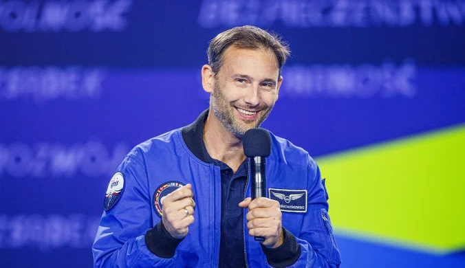 Mężczyzna w niebieskiej kurtce z emblematami astronautycznymi trzyma mikrofon i uśmiecha się podczas wystąpienia na scenie z niebieskim tłem.