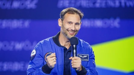 Mężczyzna w niebieskiej kurtce z emblematami astronautycznymi trzyma mikrofon i uśmiecha się podczas wystąpienia na scenie z niebieskim tłem.