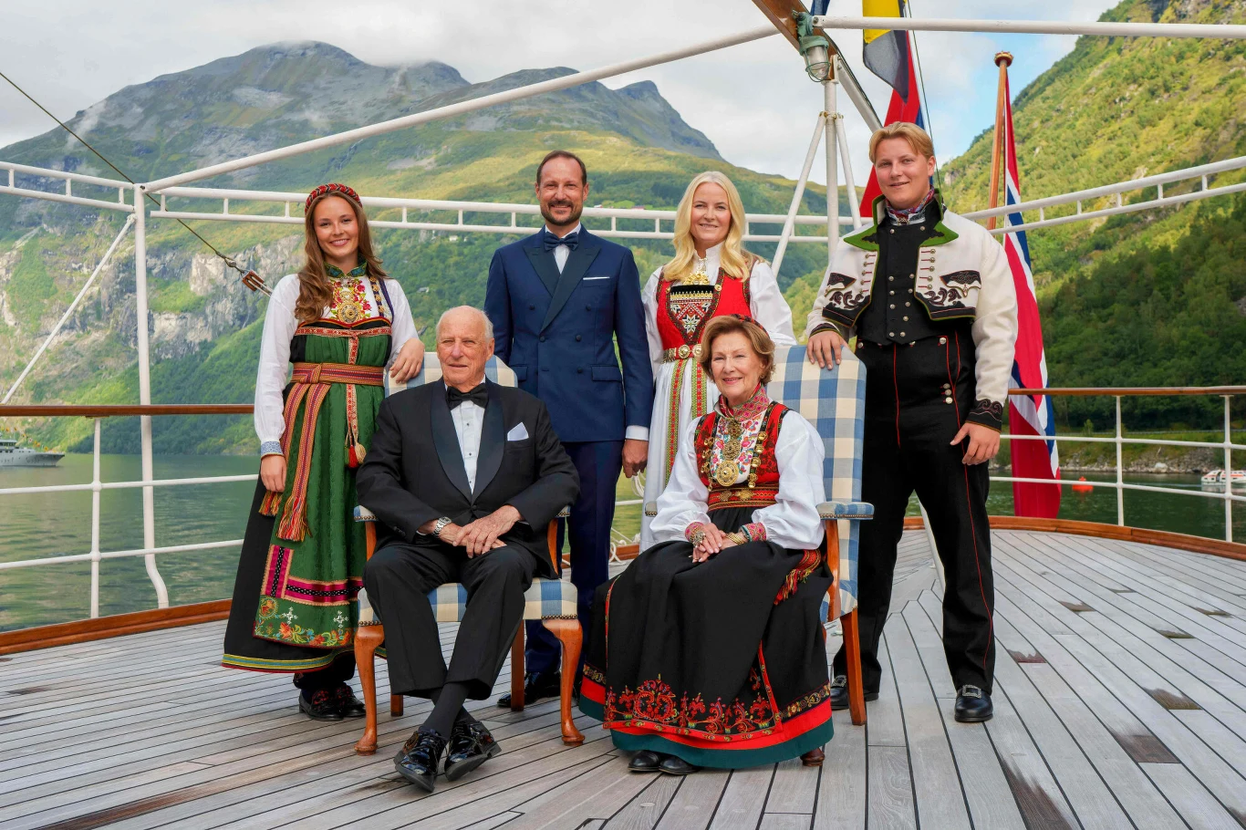 Sześć osób ubranych w tradycyjne norweskie stroje oraz garnitur, stojących i siedzących na pokładzie statku, w tle góry, norweska flaga i woda.