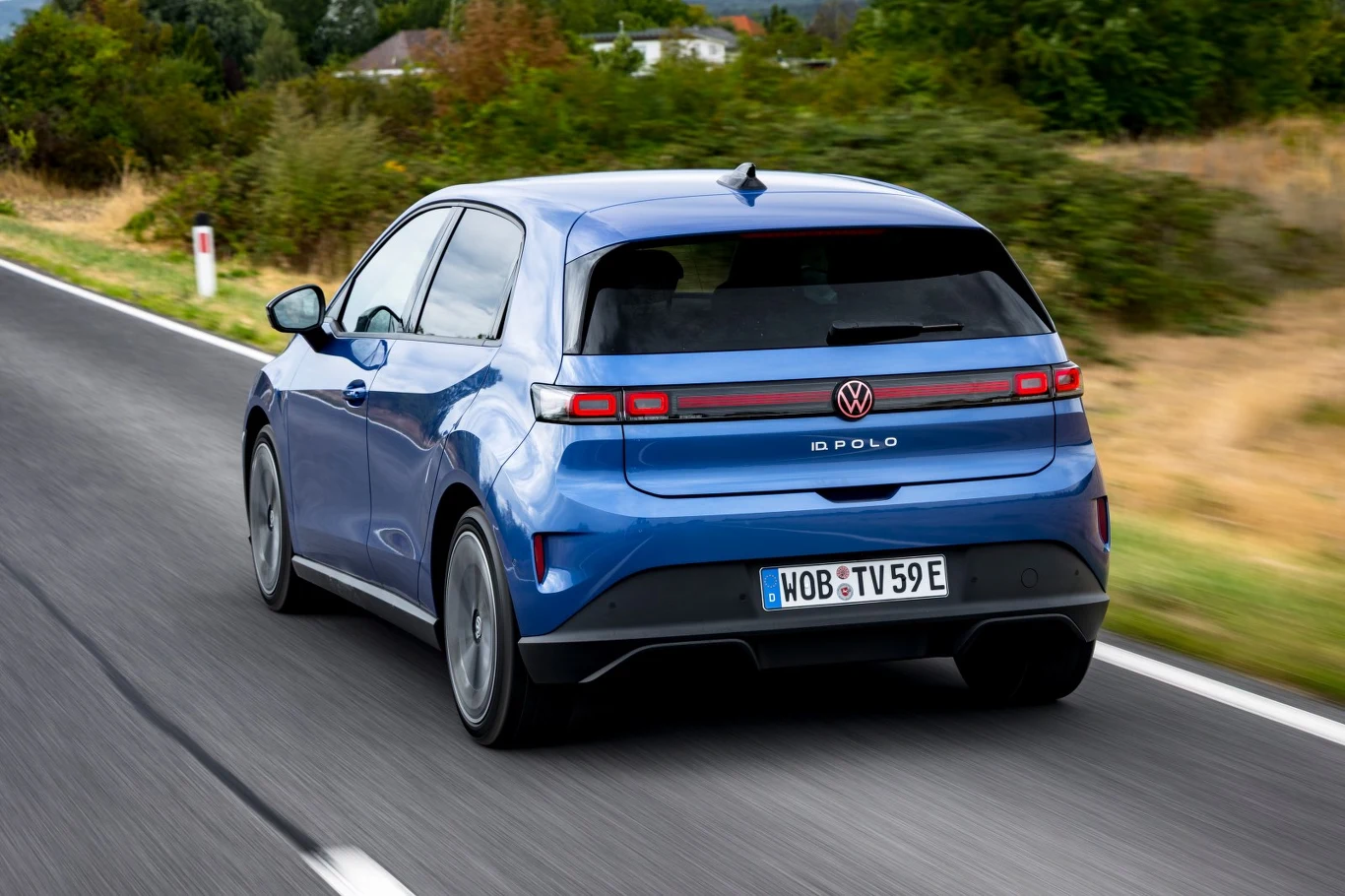 Stylizacja Volkswagena ID. Polo wyraźnie odróżnia go od jego spalinowego odpowiednika. Pod względem rozmiarów stoją blisko siebie - elektryk jest nieznacznie szerszy, wyższy i ma większy rozstaw osi, ale nadwozie jest nieco krótsze. Niebieski Volkswagen Polo jedzie asfaltową drogą otoczoną zielenią i suchą trawą.