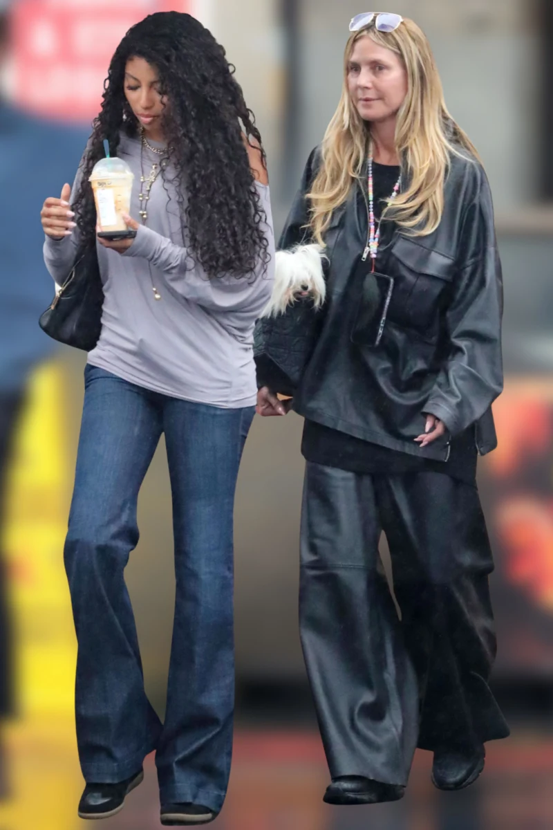 Naturalna Heidi Klum na zakupach z córką w Los Angeles Dwie kobiety spacerujące razem, jedna ubrana w szerokie dżinsy i fioletową bluzkę z odkrytym ramieniem, trzymająca napój, druga w czarnym, obszernym stroju ze sztucznego materiału i okularach na głowie, prowadząca małego białego psa na rękach.