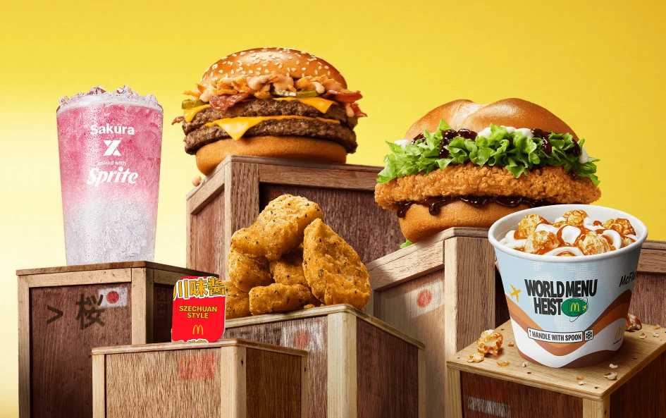 Dwuwarstwowy burger z bekonem i serem, kanapka z chrupiącym kurczakiem, różowy napój gazowany, porcja nuggetsów oraz kubełek z deserem lodowym na tle jasnych drewnianych skrzyń i żółtego gradientu.