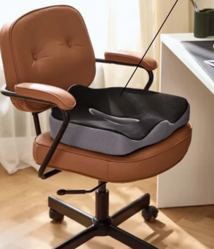 Brązowy, obrotowy fotel biurowy z podłokietnikami, na którym leży czarna, ergonomiczna poduszka wspierająca prawidłową postawę, ustawiony przy białym biurku w jasnym, domowym wnętrzu.