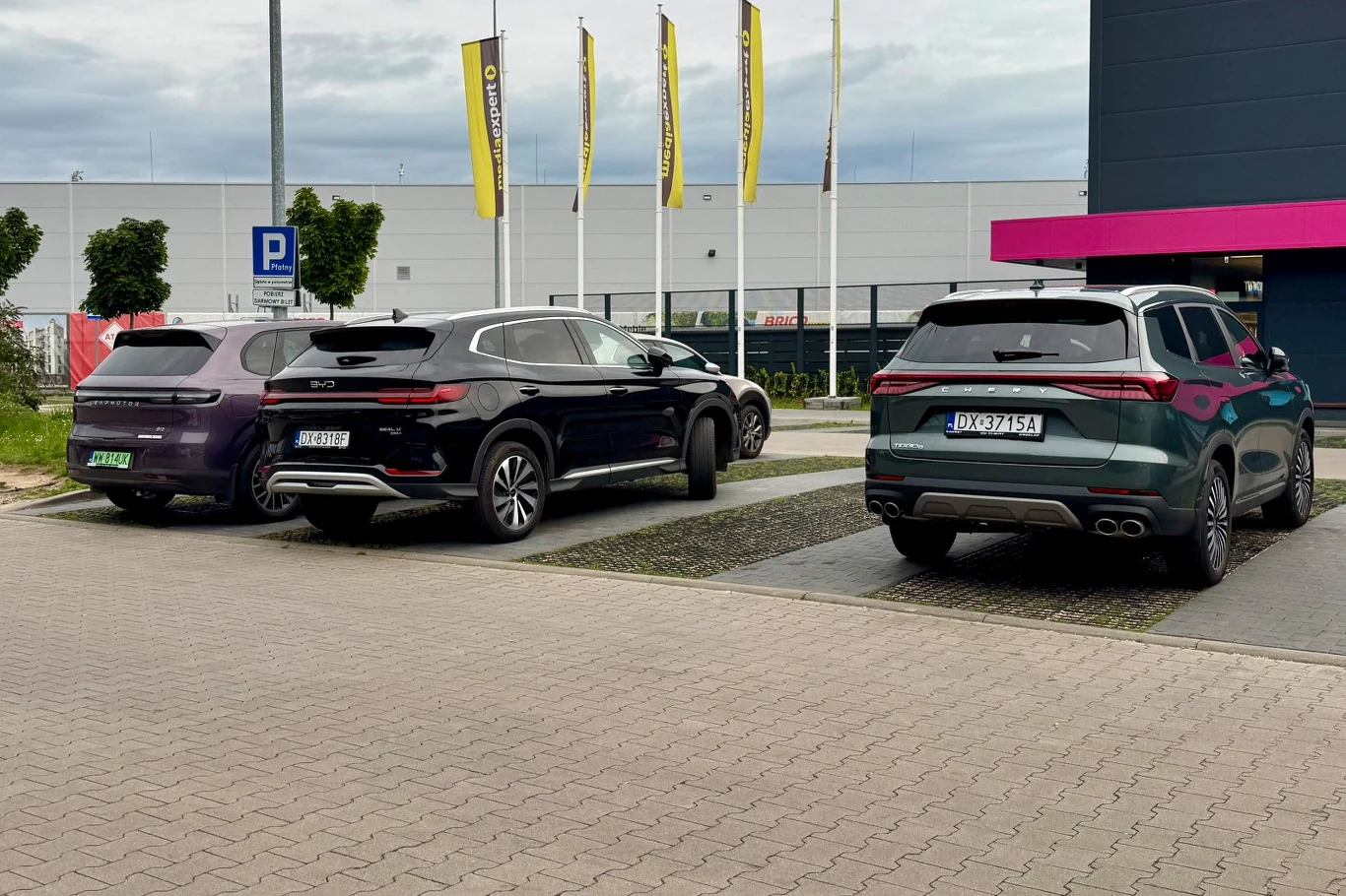 Chińskie samochody coraz częściej trafiają na listy zakupowe polskich firm. Największe obawy budzą serwis i dostępność części. Trzy chińskie SUV-y z europejskimi tablicami na parkingu, w tle flagi reklamowe i budynek.