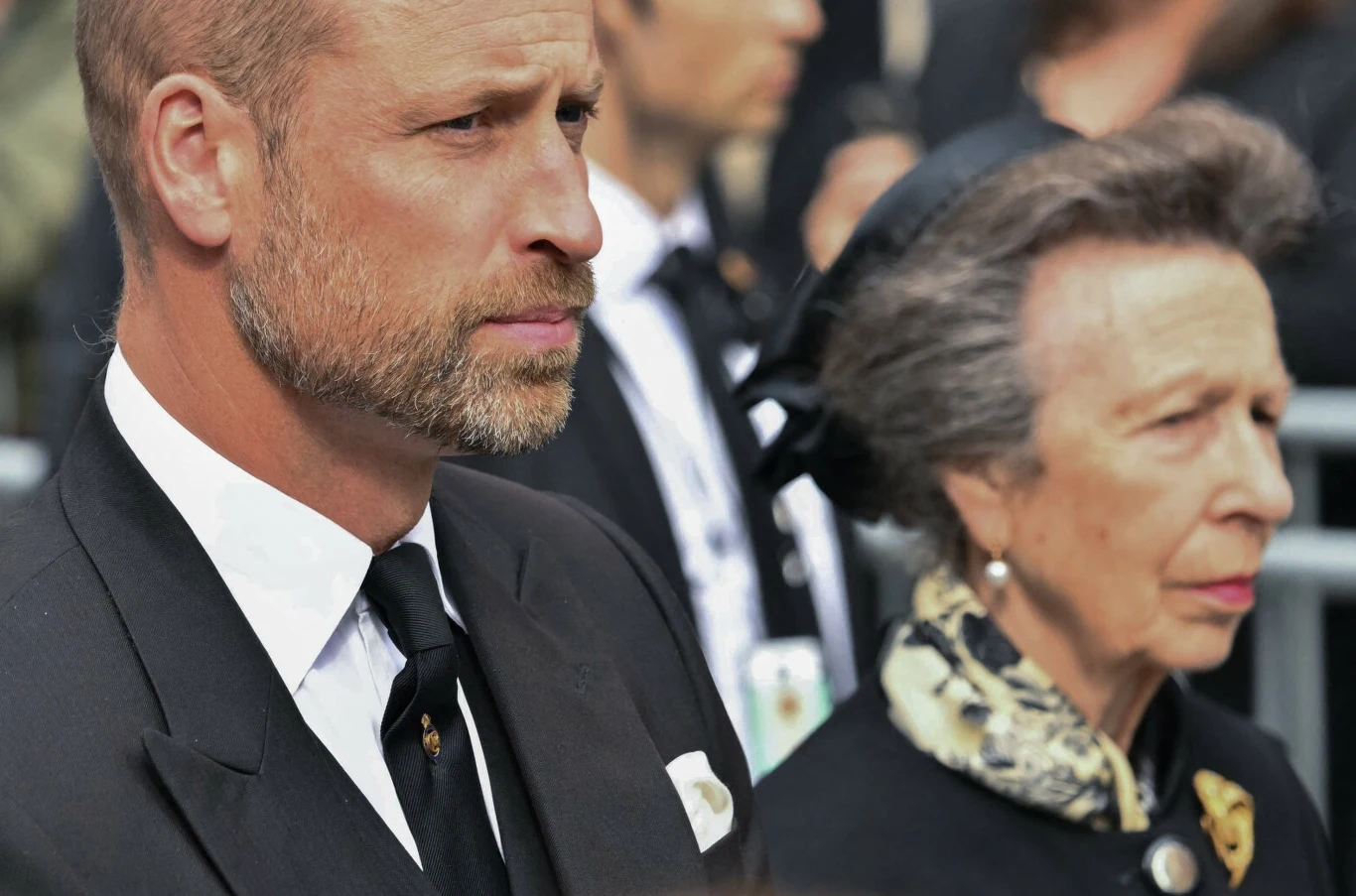 Dwie osoby w eleganckich, ciemnych strojach żałobnych stoją obok siebie z poważnym wyrazem twarzy, kobieta ma na głowie czarny kapelusz z siateczką oraz apaszkę z jasnym wzorem.