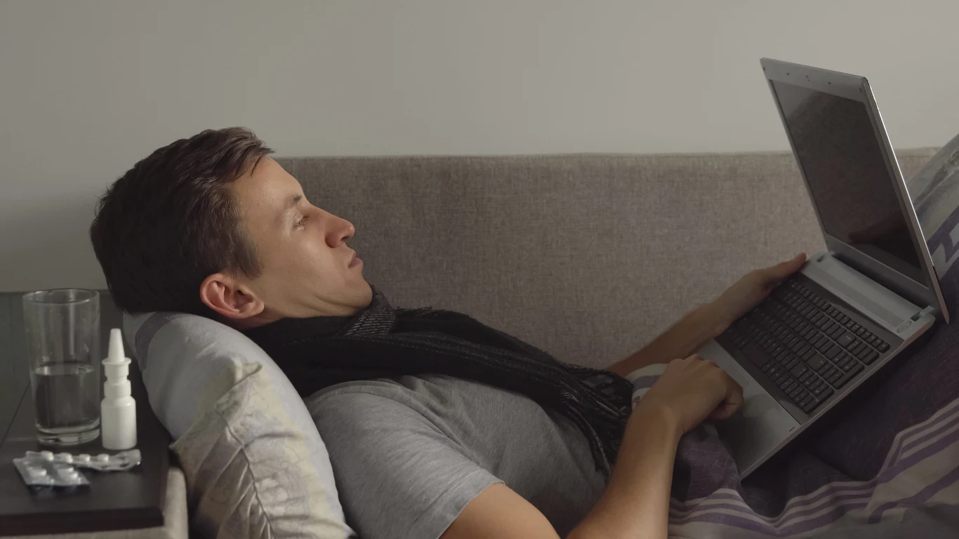 Mężczyzna leżący na kanapie pod kocem, korzystający z laptopa, obok na stoliku szklanka wody, tabletki oraz spray do nosa.