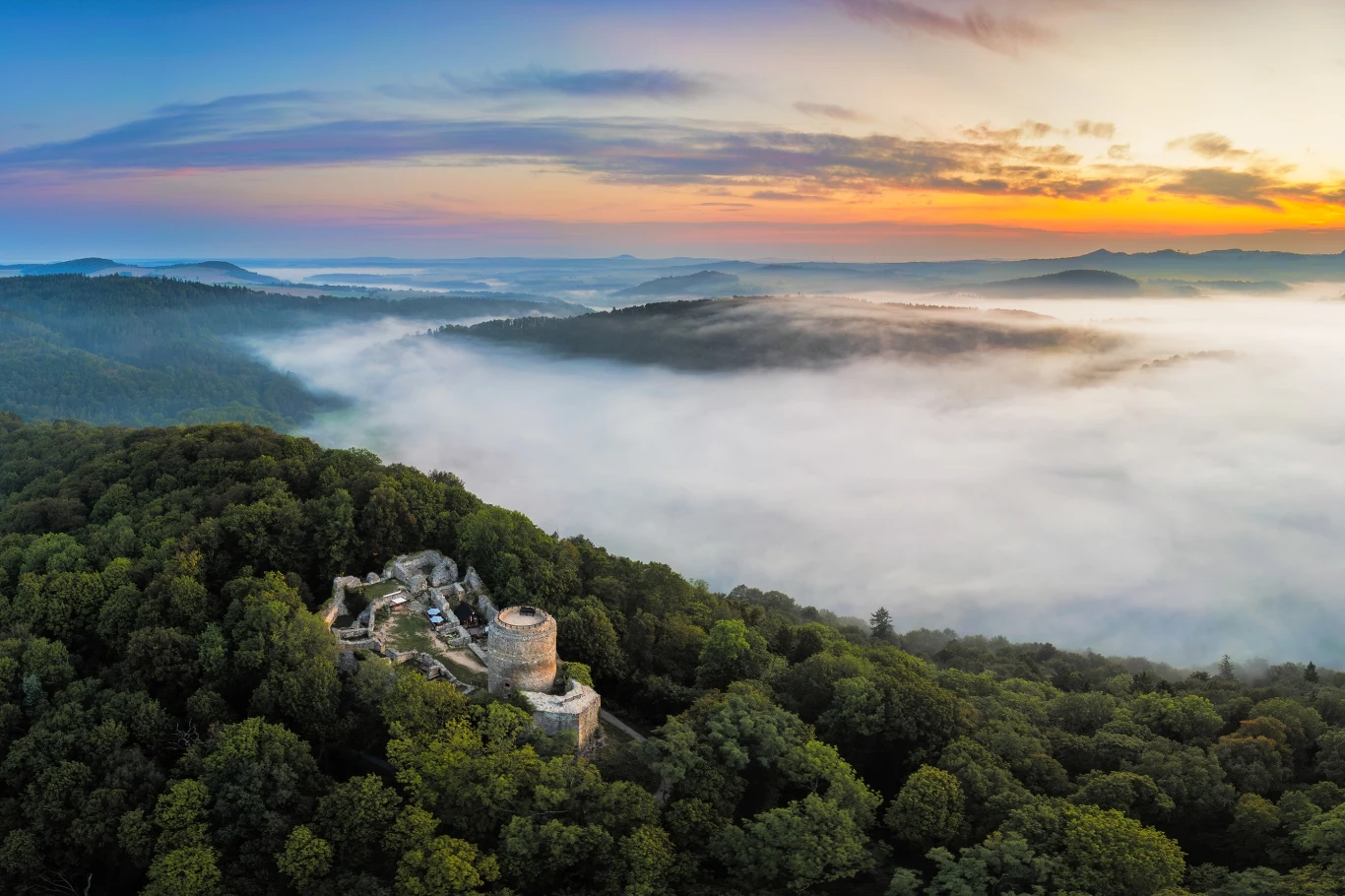 Poranna panorama zamku Wleń Ruiny średniowiecznego zamku pośród gęstego, zielonego lasu, otulone poranną mgłą, w tle rozciągające się wzgórza oraz kolorowy, wschodzący słońcem horyzont.