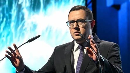 Mężczyzna w garniturze i okularach przemawia przy mównicy, gestykulując rękami, na tle jasnego, efektownego ekranu.