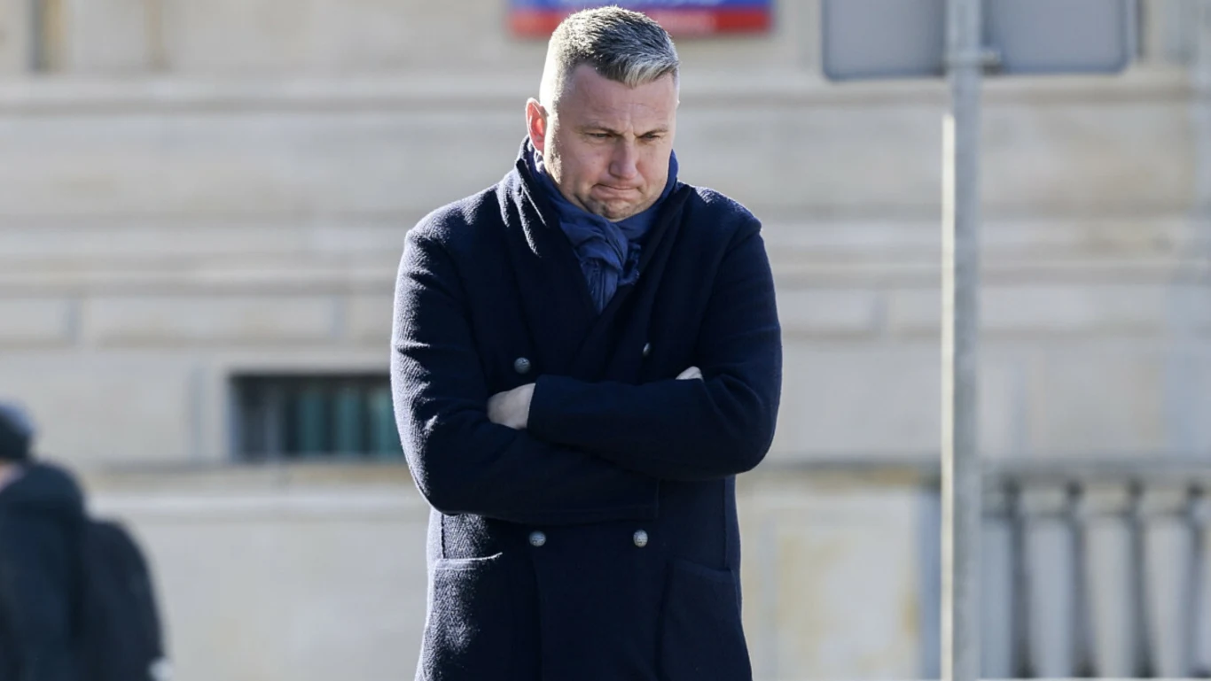 Mężczyzna w granatowym płaszczu i szaliku stoi z założonymi rękami, patrząc w dół z poważnym wyrazem twarzy, na tle jasnej ściany budynku.