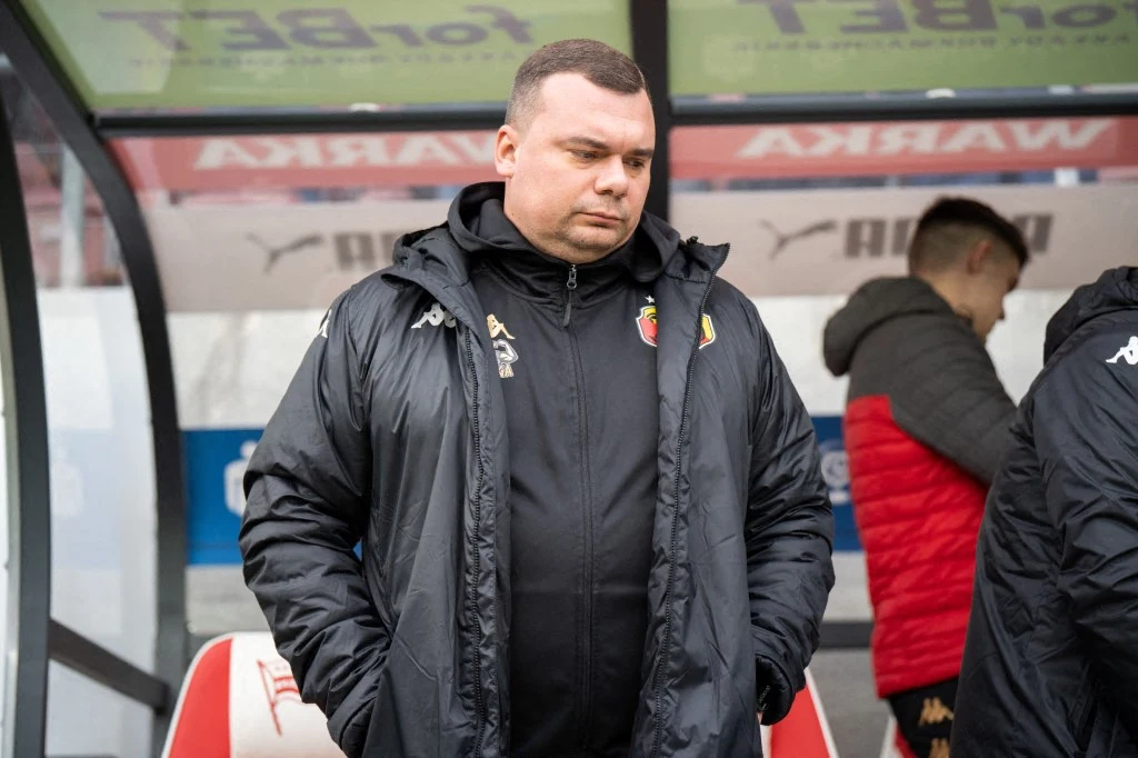 Mężczyzna w czarnej kurtce sportowej stoi przy ławce rezerwowych na stadionie piłkarskim, wyglądając poważnie i skupiony; w tle widoczny inny członek drużyny w czerwonej kurtce.