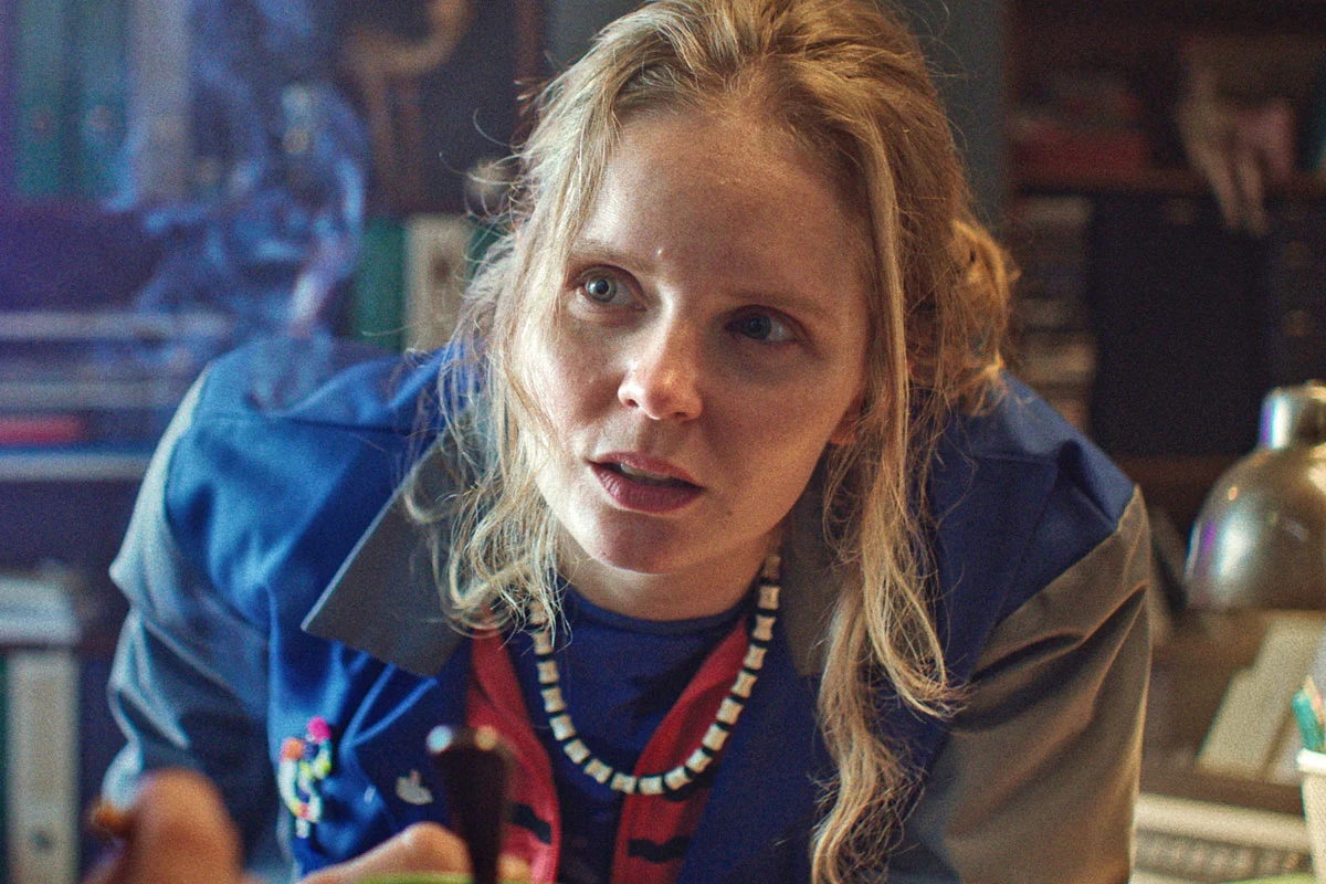 Olga Kalicka w scenie z filmu "Ma to sens" Kobieta w niebieskim uniformie pochylona nad biurkiem, w ręku trzyma czarny przedmiot, na szyi biało-czarny naszyjnik, w tle regał z dokumentami.