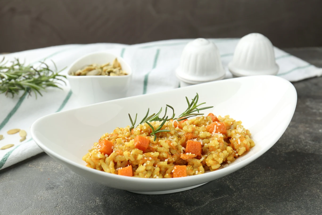 Risotto z dynią to kremowe, jesienne danie Risotto z dynią, kremowym ryżem, kawałkami dyni i gałązką rozmarynu w białej misce na szarobrązowym blacie.