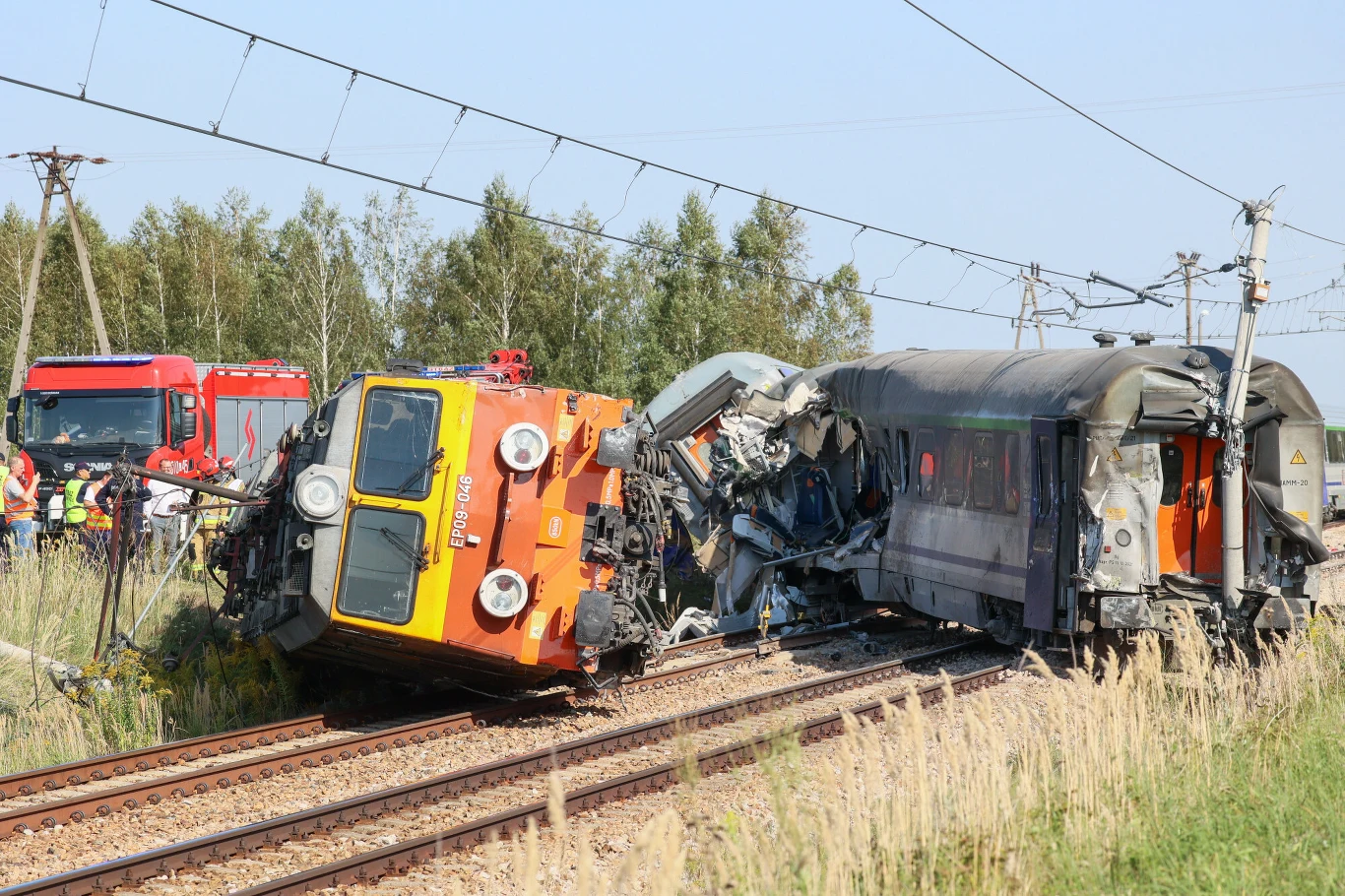 Widoczne zderzenie pociągu technicznego z pasażerskim, wykolejone wagony, poważne zniszczenia w obrębie kabiny i bocznej części składów, na miejscu obecne służby ratownicze.