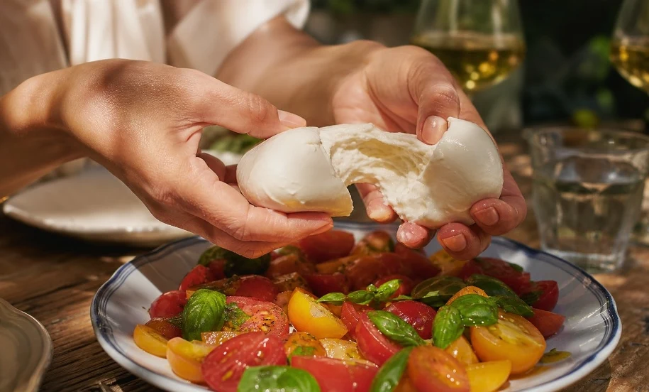 Burrata z pomidorami — letnie podanie na tarasie Dłonie rozrywają kulkę burraty nad talerzem kolorowych pomidorów z bazylią