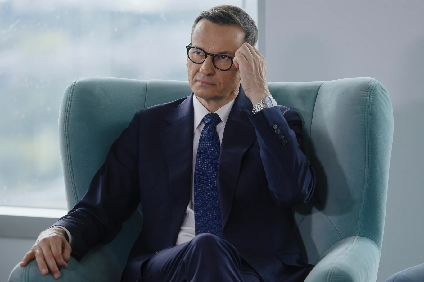 Burza po wywiadzie Morawieckiego. Poszło o Zondacrypto Mateusz Morawiecki w garniturze i okularach siedzi w fotelu, trzymając rękę przy twarzy.