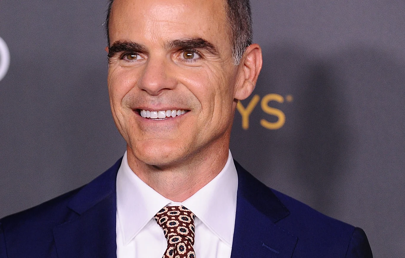 Michael Kelly Michael Kelly w granatowym garniturze, białej koszuli i wzorzystym krawacie przed ciemną ścianką z żółtym napisem