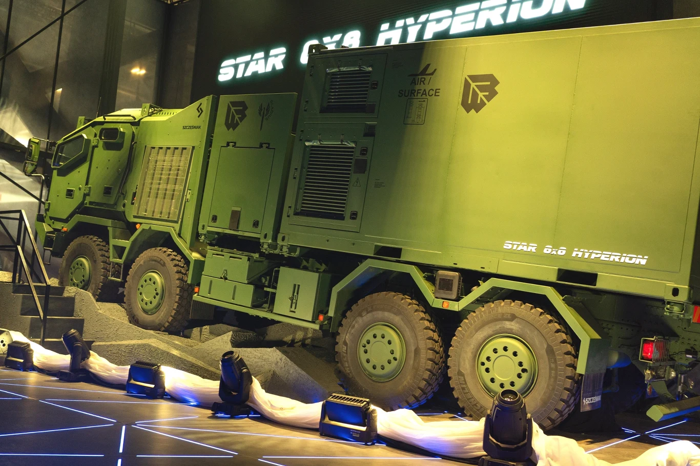 Duży wojskowy pojazd ciężarowy STAR 8x8 HYPERION w kolorze zielonym, wyposażony w liczne schowki i kratki wentylacyjne, prezentowany na podestach w otoczeniu sztucznego oświetlenia oraz napisu reklamowego na ścianie.