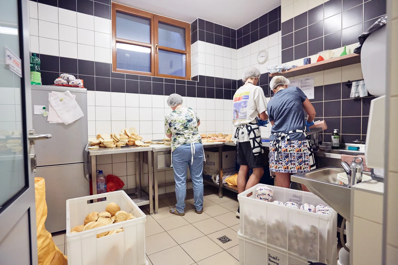 Dzieło Pomocy św. Ojca Pio Trzy osoby przygotowujące kanapki przy kuchennych blatach, wokół poukładane bochenki chleba i kosze z pieczywem.