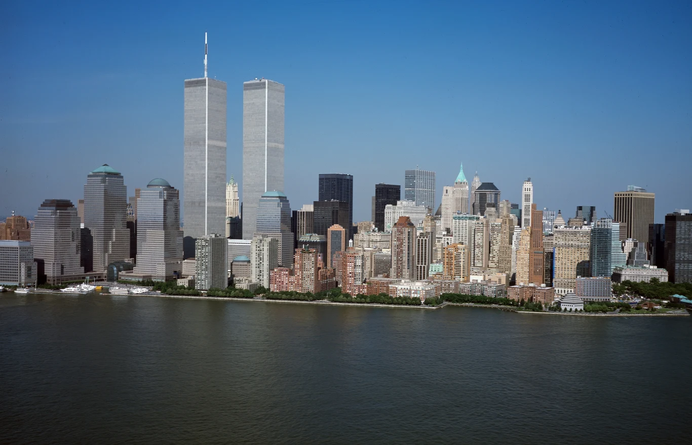 Panorama miasta z dominującymi dwoma wysokimi wieżami World Trade Center na tle licznych wieżowców, z rzeką na pierwszym planie i błękitnym niebem w tle.
