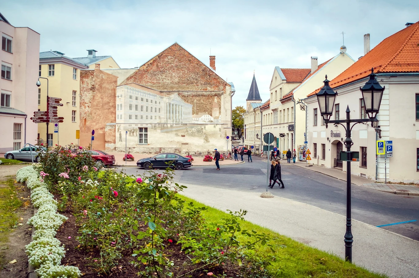 Kolorowe kamienice z ceglaną ścianą ozdobioną muralem, niewielki plac miejski z latarniami i niską roślinnością, kilka zaparkowanych samochodów oraz przechodzący piesi.
