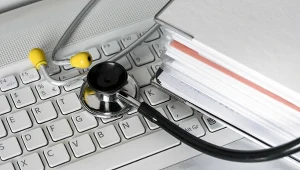 Stetoskop medyczny leży na klawiaturze laptopa obok dużego segregatora z dokumentami.