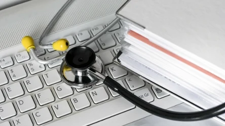 Stetoskop medyczny leży na klawiaturze laptopa obok dużego segregatora z dokumentami.