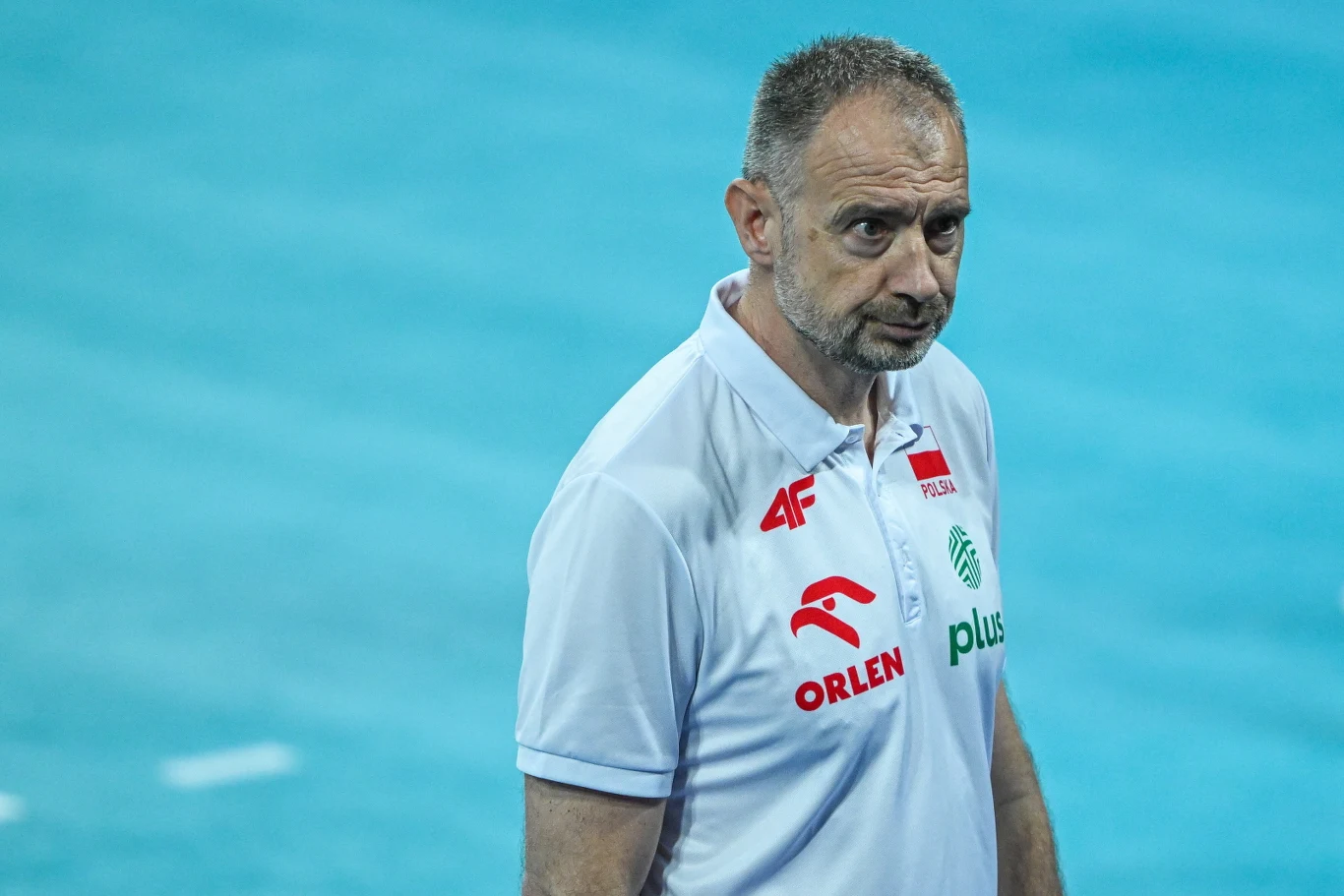 Nikola Grbić Mężczyzna w białej koszulce z logo Orlen, Plus i 4F oraz polską flagą stoi na tle niebieskiego boiska sportowego.