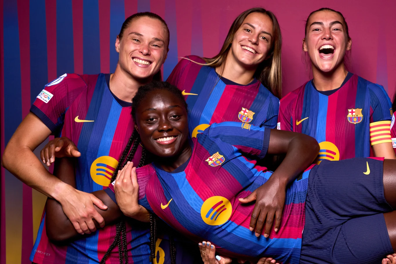Cztery piłkarki ubrane w barwy FC Barcelony uśmiechają się, trzy z nich obejmują uniesioną koleżankę, prezentując radość i sportową integrację.