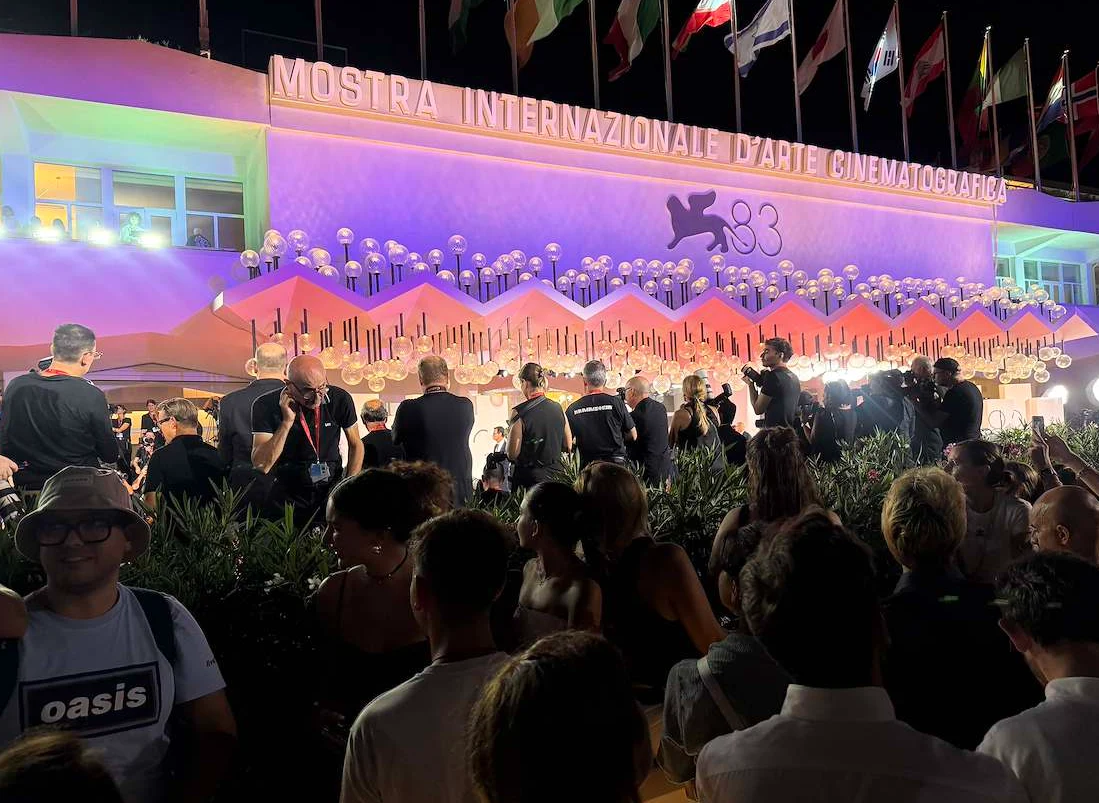 Tłum ludzi zgromadzony przed jasno oświetlonym budynkiem festiwalu filmowego w Wenecji, nad którym powiewają liczne flagi, a na fasadzie widoczny jest logotyp imprezy i napis „Mostra Internazionale d’Arte Cinematografica”.