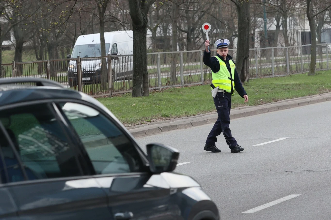 Według zapowiedzi ministra, wjazd na tory przy zapalonym czerwonym świetle będzie oznaczał utratę prawa jazdy na 3 miesiące. Policjant w żółtej kamizelce zatrzymuje samochód osobowy na ulicy za pomocą lizaka drogowego.