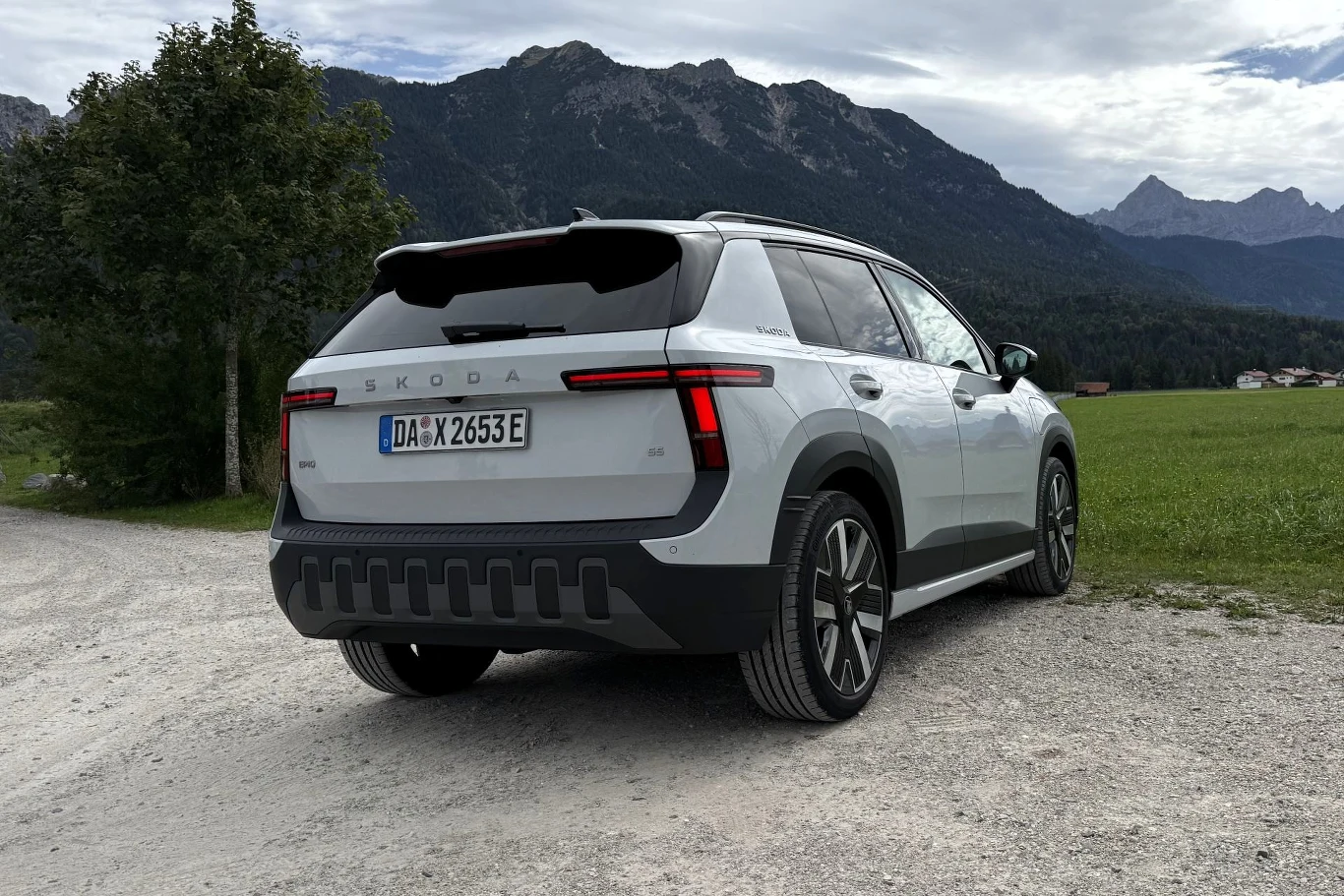Biała Skoda o nowoczesnym designie i czarnych detalach stoi zaparkowana na żwirowej drodze, w tle wysokie, zalesione góry i rozległa polana.