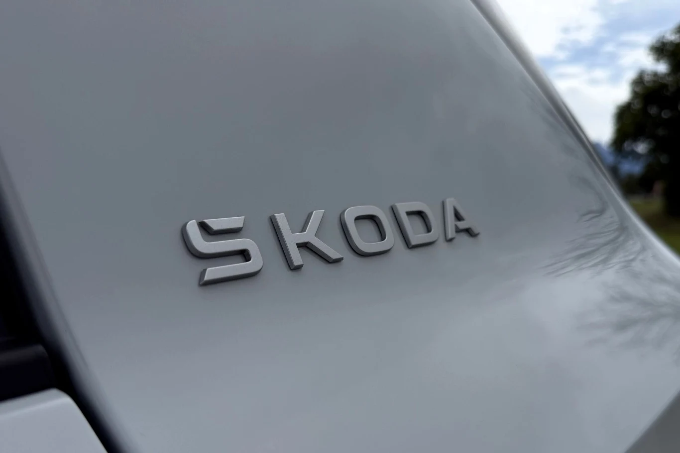 Srebrne logo marki Škoda umieszczone na jasnej karoserii samochodu.
