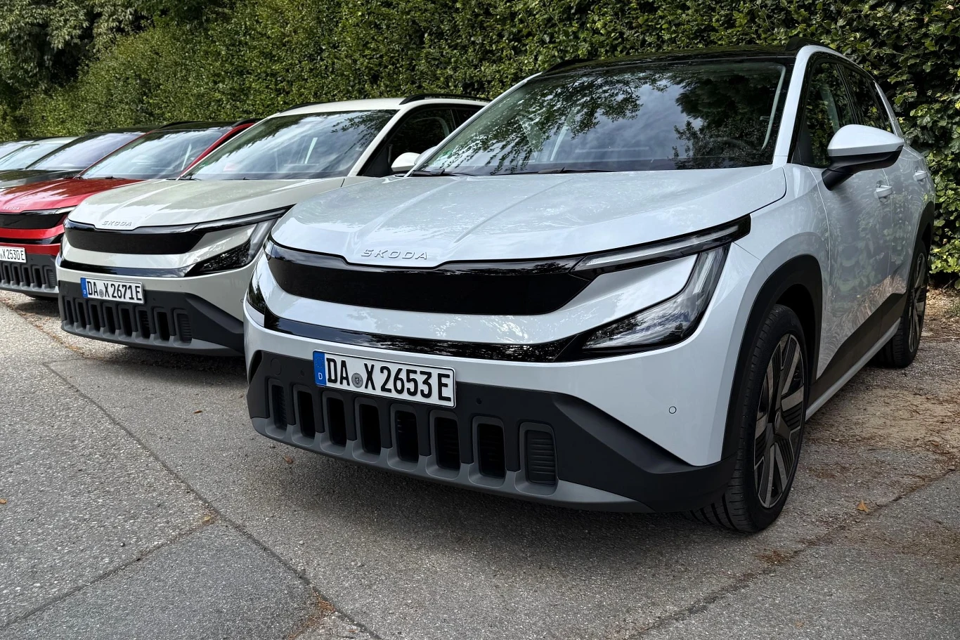 Nowoczesne SUV-y marki Skoda w jasnych kolorach ustawione w szeregu na parkingu, widoczne charakterystyczne reflektory oraz minimalistyczny design przodów pojazdów.